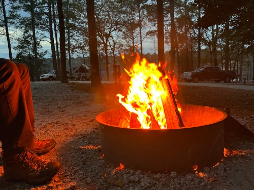 A campfire burns brightly in a fire pit, with a person's foot visible nearby, surrounded by trees at dusk.
