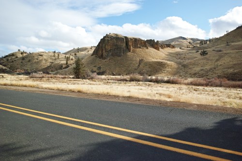 A scenic view of a road beside dry, hilly terrain with rocky formations and sparse vegetation under a partly cloudy sky.