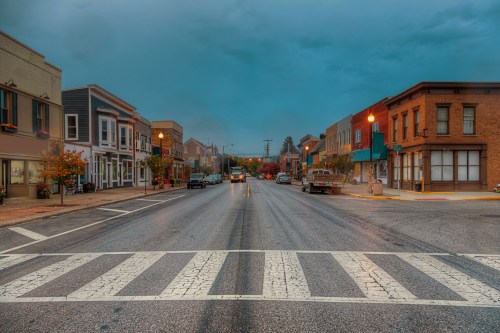 A quiet street scene with shops on either side, under a cloudy sky, and a crosswalk in the foreground.