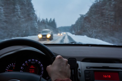 A driver's hand on the steering wheel, navigating a snowy road with oncoming headlights in the distance.
