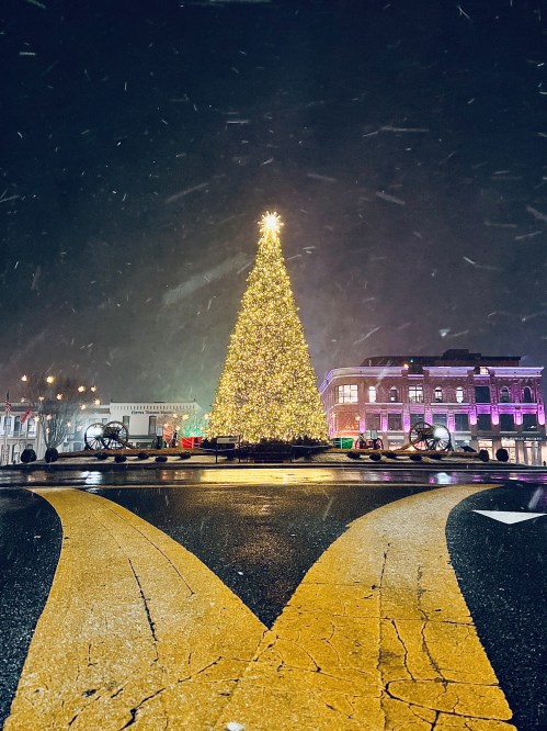 A brightly lit Christmas tree stands in the center of a snowy street, with festive buildings in the background.
