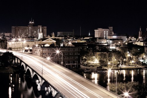 Nighttime cityscape featuring a bridge, illuminated buildings, and reflections on the water.