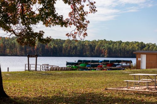 A serene lakeside scene with colorful kayaks stacked by the water, surrounded by trees and a small building.