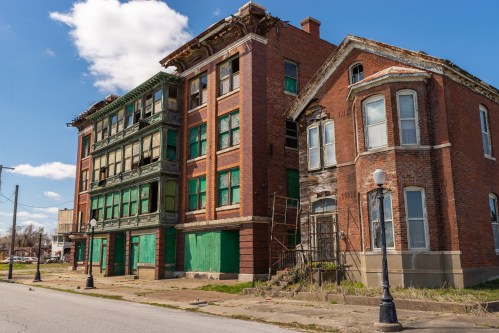 Abandoned brick buildings with boarded windows and peeling paint under a blue sky.