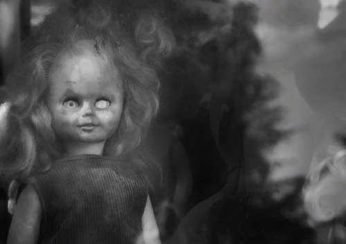 A close-up of a vintage doll with a cracked face and wild hair, set against a blurred, dark background.