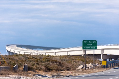 A curved concrete bridge with a green sign for Marc Basnight Bridge and Oregon Inlet against a cloudy sky.