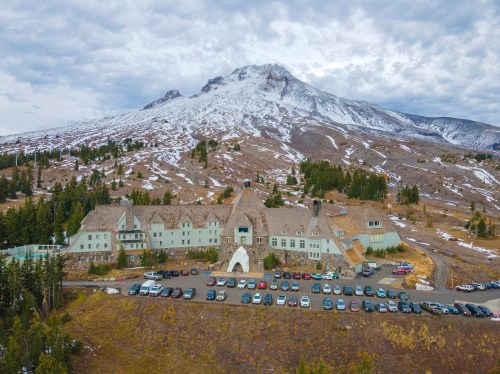 Aerial view of a large lodge with a snowy mountain backdrop and a parking lot filled with cars.