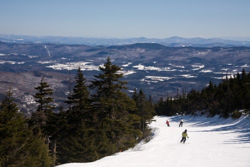 Skiers descend a snowy slope with a panoramic view of mountains and valleys under a clear blue sky.