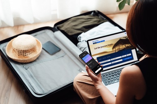 A person sitting beside an open suitcase, using a laptop and smartphone, planning a trip with clothes and a hat nearby.