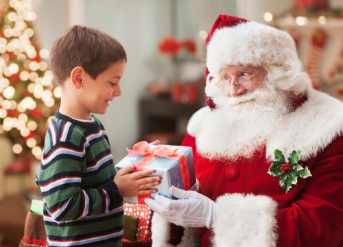 A young boy smiles as he receives a gift from Santa Claus in a festive setting with a Christmas tree in the background.