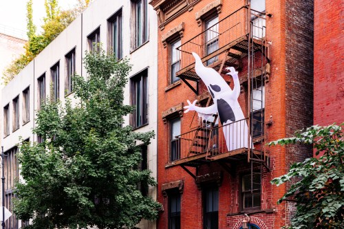 A large ghost decoration hangs from a fire escape on a red brick building, surrounded by greenery and other buildings.