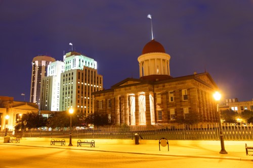 Historic building with a red dome, illuminated at night, alongside modern skyscrapers and street lamps.