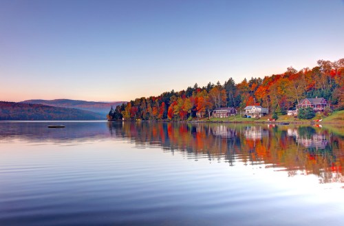 A serene lake at dawn, reflecting colorful autumn trees and charming houses along the shore.
