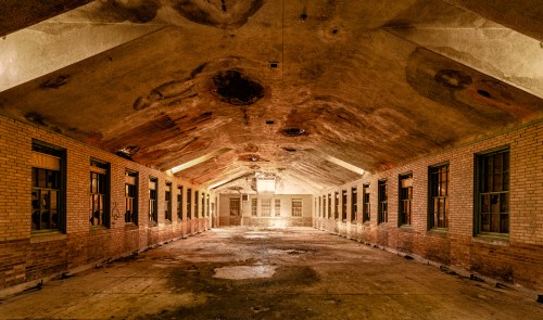 Abandoned interior of a long, narrow room with brick walls, a high ceiling, and water damage, illuminated by soft light.