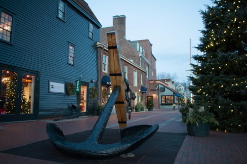 A large anchor sculpture in a brick-paved street, flanked by shops and a decorated tree during twilight.