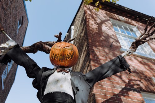 A scarecrow with a carved pumpkin head stands against a brick wall, arms outstretched, wearing a tattered suit.