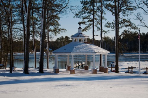 A snow-covered gazebo by a frozen lake, surrounded by tall pine trees under a clear blue sky.