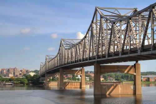 A steel bridge spans a river, with a city skyline visible in the background under a clear blue sky.