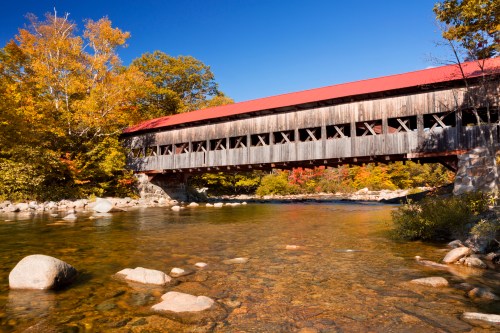 A scenic view of a covered bridge over a clear river, surrounded by colorful autumn foliage and a blue sky.