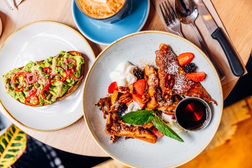 A plate of French toast topped with berries and mint, alongside avocado toast with red pepper slices and a coffee.