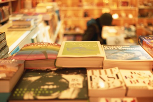 A stack of books on a table in a cozy bookstore, with a blurred figure browsing in the background.