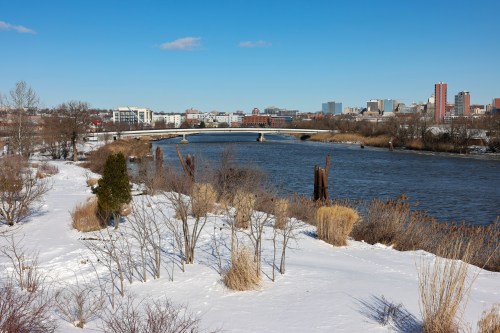 A snowy riverside scene with a bridge, city skyline, and bare trees under a clear blue sky.