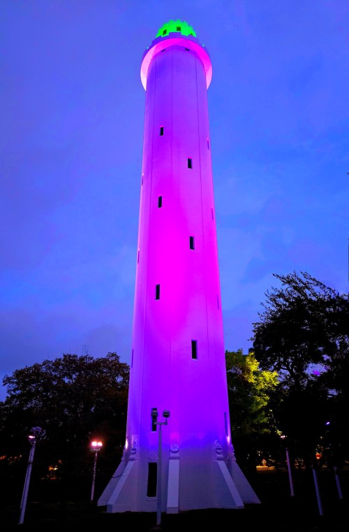 A tall, illuminated tower glows in purple and green against a twilight sky, surrounded by trees and soft lighting.