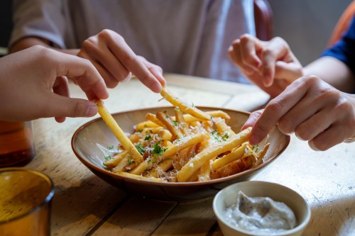 Hands reaching for a bowl of fries topped with herbs, alongside a small dish of dipping sauce on a wooden table.