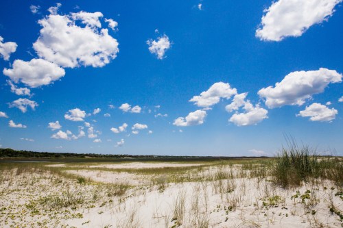 A sandy beach with green grass and a clear blue sky dotted with fluffy white clouds.