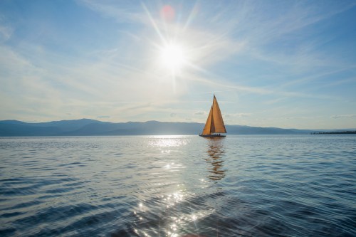 A sailboat glides across a shimmering lake under a bright sun and clear blue sky, with mountains in the background.