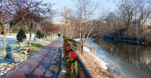 A scenic riverside path lined with trees, benches, and festive decorations, under a clear blue sky. Snow on the ground.