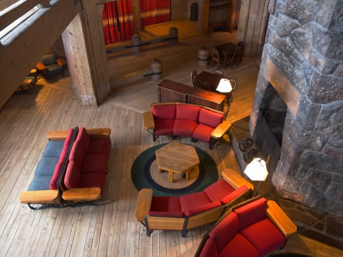 Cozy lodge interior with wooden floors, circular rug, and red upholstered seating around a central table and fireplace.