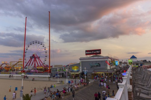 A lively boardwalk scene at sunset, featuring a ferris wheel, amusement rides, and crowds of people enjoying the atmosphere.