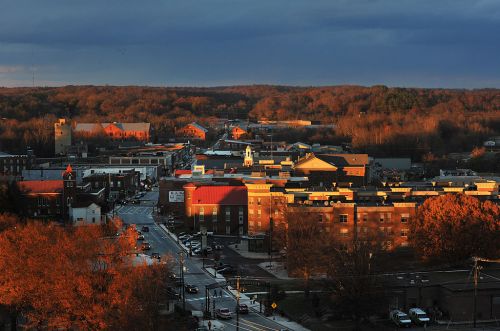 Aerial view of a small town with autumn foliage, featuring buildings and streets under a partly cloudy sky.