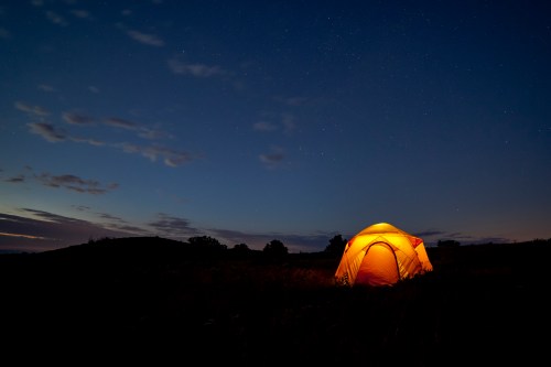 A glowing tent in a field under a starry night sky, with soft clouds and a hint of twilight.