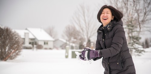 A woman in a winter coat joyfully throws snow in a snowy landscape, smiling with a house in the background.