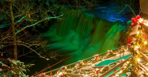 A snowy scene with a waterfall illuminated in green, surrounded by festive lights and decorations on a wooden railing.
