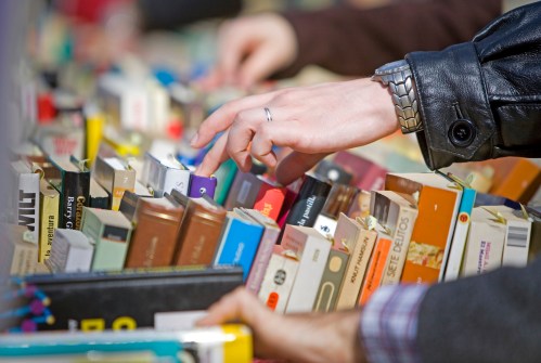 A hand reaches for a book among a colorful array of books stacked on a table.