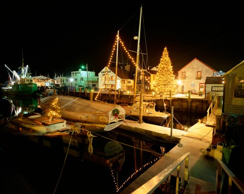 A festive harbor at night, featuring decorated boats, a Christmas tree, and illuminated buildings along the water.