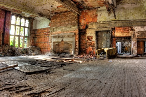 Abandoned room with crumbling walls, debris on the floor, and a dilapidated fireplace, illuminated by natural light.