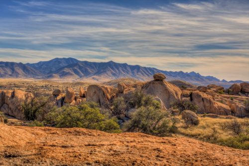 A scenic landscape featuring large boulders, rolling hills, and distant mountains under a blue sky with wispy clouds.