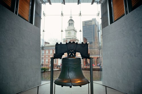 The Liberty Bell stands in a modern museum, with a historic building and clock tower visible through glass behind it.