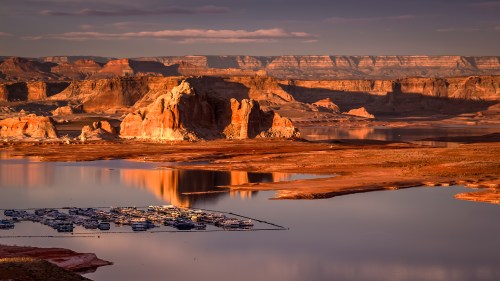A serene landscape featuring a lake with a marina, surrounded by rocky cliffs and mountains at sunset.