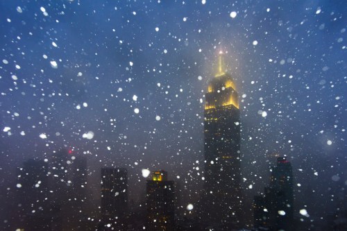 Snow falls heavily over a city skyline, with the illuminated Empire State Building visible in the background.