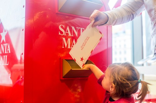 A child reaches into a red mailbox labeled "Santa Mail" while an adult drops in a letter.