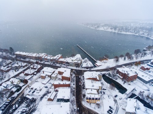 Aerial view of a snowy town by a lake, with snow-covered rooftops and a pier extending into the water.