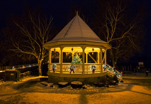 A snow-covered gazebo at night, adorned with festive lights and a small Christmas tree inside.