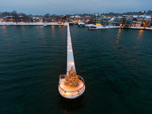 A snowy pier extends into a lake, featuring a lit Christmas tree at its end, with buildings visible in the background.