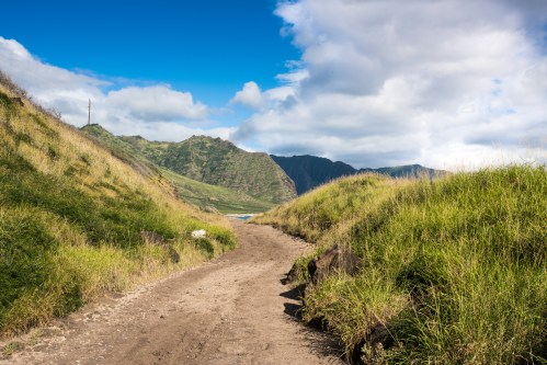 A sandy path winds through lush green hills, leading to a scenic view of mountains and a blue sky.