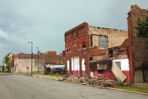 Abandoned buildings with crumbling brick facades and debris along a deserted street under a cloudy sky.